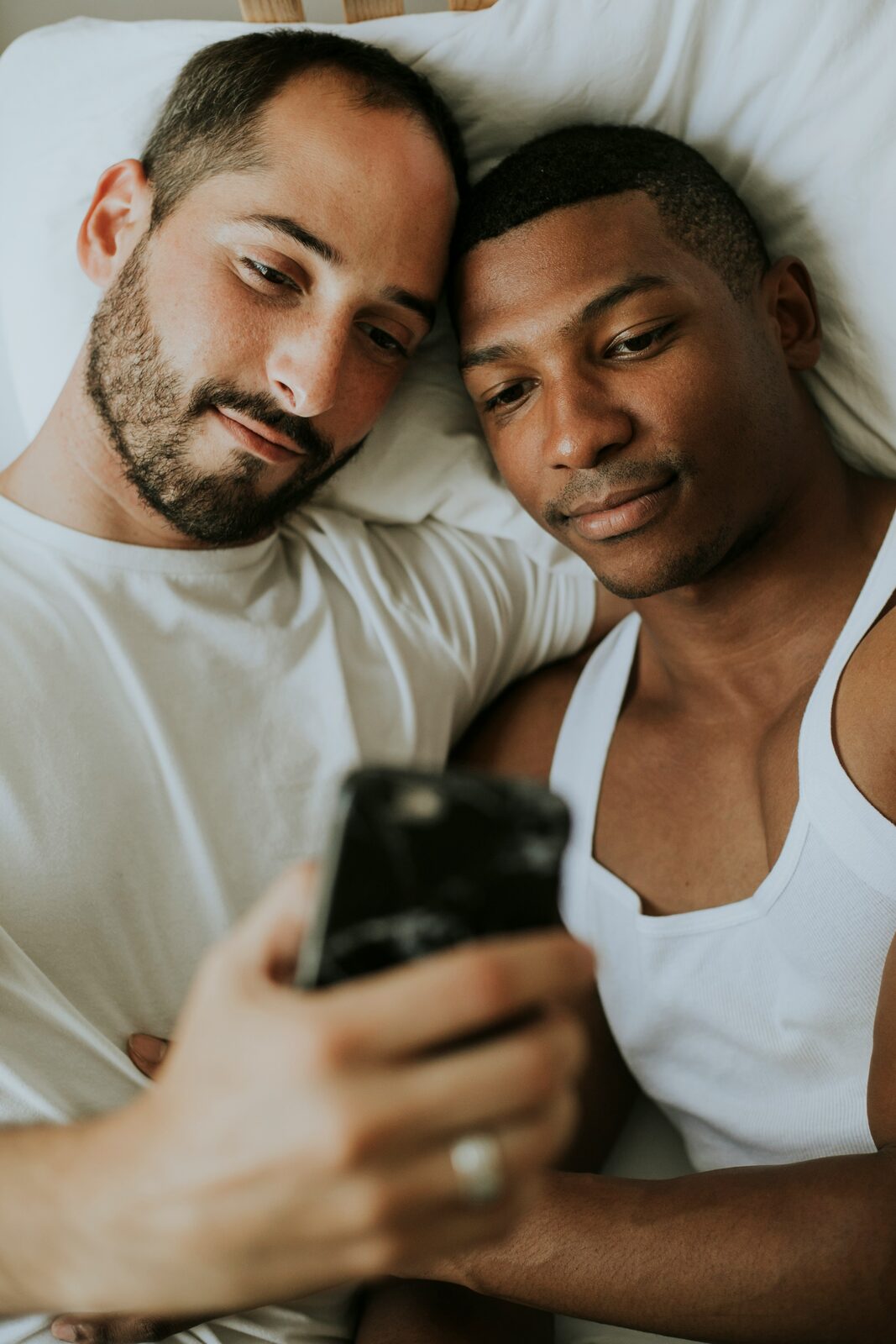 Two men lying in bed together, looking at a phone.